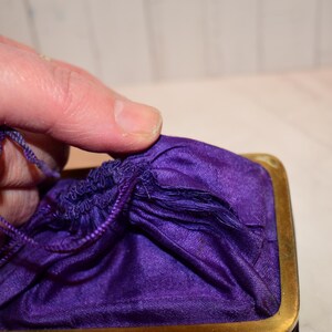 May include: A close-up of a small, vintage purple satin drawstring bag with a gold-toned metal frame. The bag is partially open, revealing the gathered fabric and drawstring closure. The bag is held by a hand.