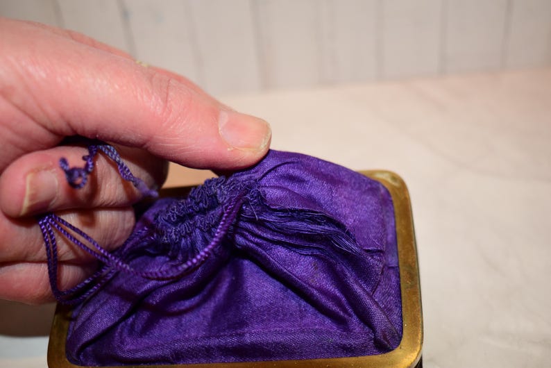 May include: A close-up of a small, vintage purple satin drawstring bag with a gold-toned metal frame. The bag is partially open, revealing the gathered fabric and drawstring closure. The bag is held by a hand.