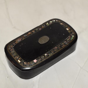 May include: A rectangular black box with a decorative border of iridescent shell pieces. The box has a small, oval, silver-colored accent on the top. The box is set against a white marble surface.