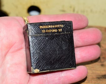 May include: A small, black, rectangular box with gold lettering that reads "PARKINS & GOTTO" and "25 OXFORD ST". The box has a textured surface and a gold-coloured loop attached to the top.