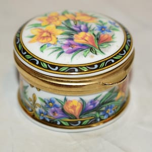 May include: A small, round, porcelain trinket box with a floral design. The lid and sides feature colorful flowers in yellow, purple, and orange, set against a white background. The box has a gold-colored metal rim and clasp.