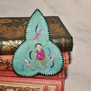 May include: A teal blue fabric applique with a black scalloped edge. The applique features a hand-stitched design of a person in a pink and yellow outfit holding a yellow object. The person is surrounded by pink and blue flowers.