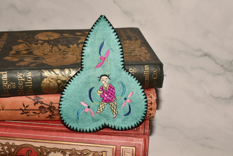 May include: A teal blue fabric applique with a black scalloped edge. The applique features a hand-stitched design of a person in a pink and yellow outfit holding a yellow object. The person is surrounded by pink and blue flowers.