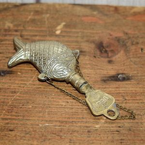 May include: An antique brass fish-shaped bottle opener with intricate engraved details. The opener has a chain attached to a rectangular tag that reads "DELHI". The opener is lying on a wooden surface.