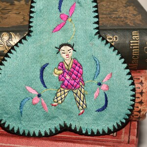 May include: A teal blue fabric heart-shaped pincushion with black trim. The pincushion is embroidered with a figure in pink and yellow clothing, holding a small object. The figure is surrounded by pink and blue flowers.