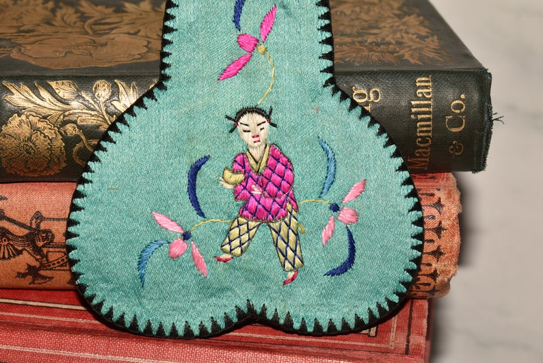 May include: A teal blue fabric heart-shaped pincushion with black trim. The pincushion is embroidered with a figure in pink and yellow clothing, holding a small object. The figure is surrounded by pink and blue flowers.