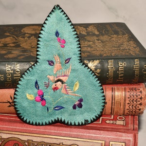 May include: A teal blue fabric bookmark with a black border and embroidered flowers and a bird. The bookmark is shaped like a teardrop and is resting on top of a stack of books.