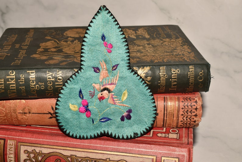 May include: A teal blue fabric bookmark with a black border and embroidered flowers and a bird. The bookmark is shaped like a teardrop and is resting on top of a stack of books.