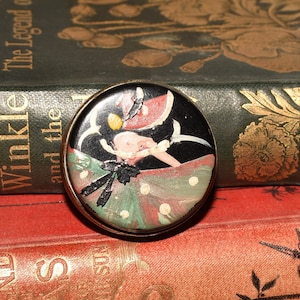 Op de afbeelding: Een vintage compact spiegel met een zwart, groen en rood ontwerp met een gestileerde vrouw in een jurk. De spiegel is rond en heeft een gouden rand.