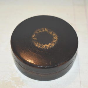 May include: A round, dark brown box with a lid. The lid features a gold-colored, circular design in the center. The box appears to be made of a smooth material, possibly leather or a similar finish.