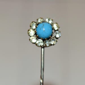 May include: A vintage hat pin with a light blue center stone surrounded by clear, sparkling rhinestones. The pin's head is flower-shaped, and the metal stem is silver-toned. This antique accessory adds a touch of elegance.