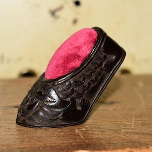 May include: A dark brown carved wooden pincushion shaped like a shoe, featuring intricate relief patterns of scales and fins across the exterior surface. The interior of the shoe is filled with a vibrant, plush, bright pink velvet cushion.