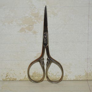 Puede incluir: Primer plano de unas tijeras de costura plateadas vintage. Las tijeras tienen una punta afilada y mangos ornamentados. Las hojas están cerradas y las tijeras están centradas sobre un fondo de papel rayado. Adecuadas para bordados o trabajos de detalle.