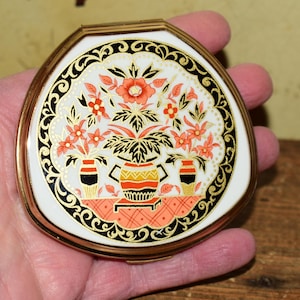 Può includere: Un compatto vintage con cornice dorata e coperchio bianco. Il coperchio presenta un motivo floreale con fiori arancioni, dettagli neri e un bordo dorato. Il compatto è tenuto in una mano.