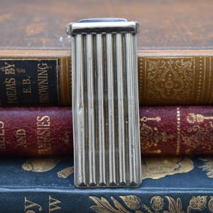 Puede incluir: Pinza para billetes de plata con un diseño acanalado. La pinza está colocada encima de una pila de libros.