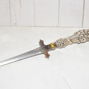 May include: A silver-toned metal letter opener featuring a pointed, tapered blade and a highly ornate, embossed handle with intricate scrollwork patterns. A small, smooth, circular brass-colored accent piece connects the blade to the handle.