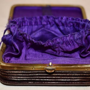 May include: An open antique purse with a dark brown exterior and a gold-coloured frame. The interior is lined with a vibrant purple fabric, and a matching drawstring pouch is visible inside. The purse is likely from the early 20th century.