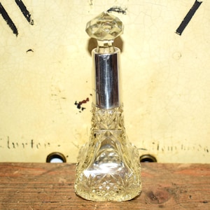 May include: A clear glass perfume bottle with a faceted, diamond-shaped stopper and a silver-colored metal neck. The bottle has a cut-glass design and sits on a wooden surface. The background is a cream-colored wall.