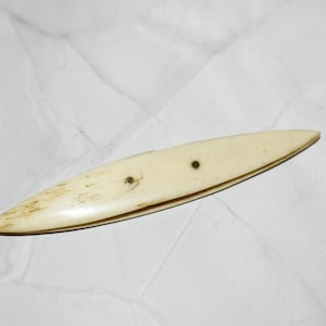 Könnte beinhalten: Ein cremefarbener, vintage Knochenfalzer. Das Werkzeug ist oval mit spitzen Enden und zwei kleinen, dunklen Nieten. Es hat ein leicht gebogenes Design und weist Alterserscheinungen mit dezenten braunen Markierungen auf. Dieses Werkzeug wird wahrscheinlich zum Glätten von Papier oder Stoff verwendet.