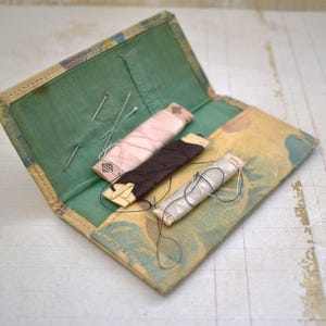 Puede incluir: Un kit de costura vintage con una cubierta de tela con estampado floral. El kit contiene tres carretes de hilo, dos de color rosa y uno marrón, y varias agujas.