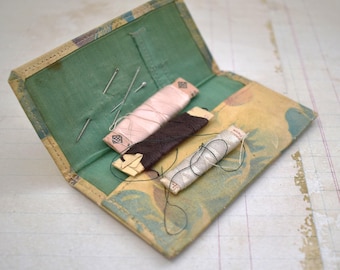 May include: A vintage sewing kit with a floral patterned fabric cover. The kit contains three spools of thread, two in pink and one in brown, and several needles.