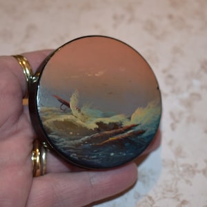 May include: A circular black compact mirror featuring a painted seascape. The artwork shows a turbulent ocean with breaking waves, a red fishing lure, and a sunset sky. The compact is held in a hand.