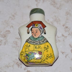 Op de afbeelding: Kleine, crèmekleurige snuiffles van keramiek met een groene stopper. De fles heeft een handgeschilderde afbeelding van een vrouw in traditionele Chinese kledij, met een gele jurk en een rode hoofdtooi. Het kunstwerk is gedetailleerd en kleurrijk.