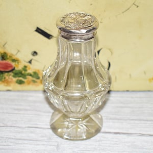 May include: A clear glass bottle with a faceted, bulbous body and a star-cut base. The bottle features a round, polished silver lid embossed with intricate floral and cherub-like motifs, providing a decorative contrast to the transparent glass.