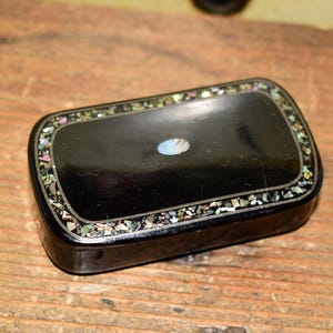 May include: A rectangular black box with rounded corners featuring a smooth, polished finish. The lid is decorated with a border of small, multicoloured crushed stone inlays and a single, centred oval-shaped iridescent shell inlay.