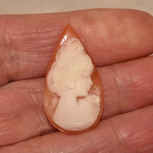 以下が含まれることがあります： 涙の形をしたカメオペンダント。女性の横顔が描かれ、髪や衣装が細かく表現されています。カメオは白で、暖かくオレンジがかった茶色の背景に映えます。ペンダントは手で持たれています。