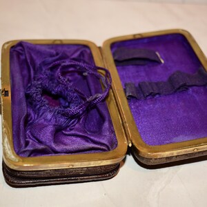 May include: An open antique case with a brass frame and a dark brown exterior. The interior is lined with purple fabric. A small drawstring bag and other items are inside. The case is on a white surface.