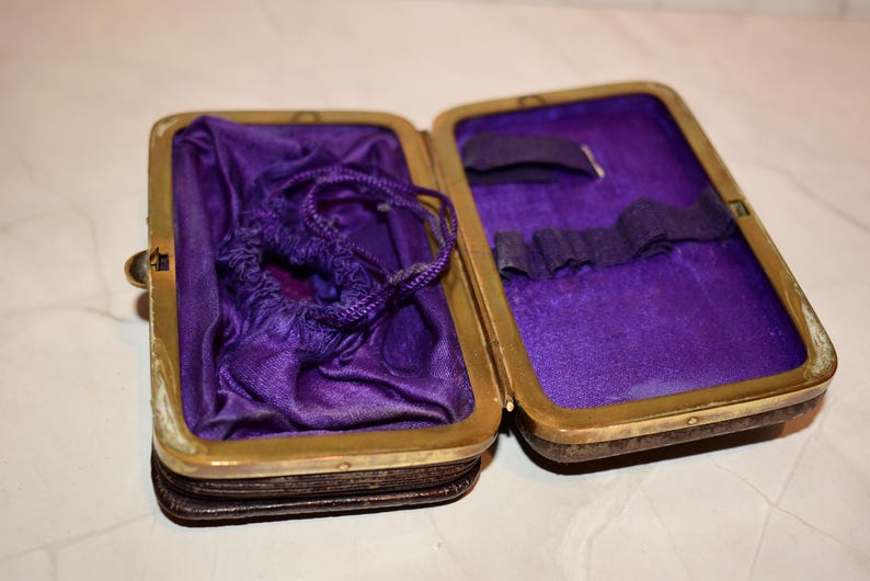 May include: An open antique case with a brass frame and a dark brown exterior. The interior is lined with purple fabric. A small drawstring bag and other items are inside. The case is on a white surface.