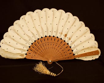 May include: An open, antique hand fan with a scalloped edge. The fan features a light brown wooden base with intricate cut-out designs and off-white, scalloped paper leaves. Small black dots are evenly spaced across the paper. A tassel hangs from the base.