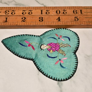 May include: A teal blue fabric patch with a black stitched border. The patch features an embroidered design of a person in pink and yellow clothing with blue and pink flowers.
