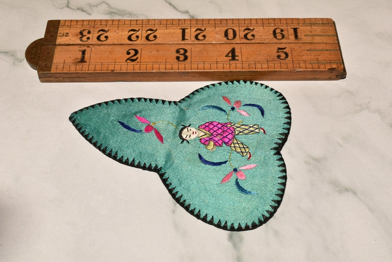 May include: A teal blue fabric patch with a black stitched border. The patch features an embroidered design of a person in pink and yellow clothing with blue and pink flowers.