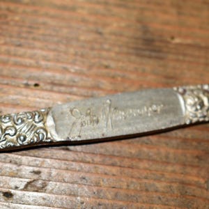 May include: A silver-toned metal letter opener featuring an ornate, embossed scrollwork pattern on the handle. The centre of the handle is engraved with the cursive text John Newspaper and the left side displays the text T D 1-94.