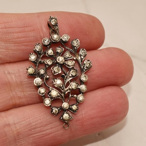 Puede incluir: Colgante plateado con diseño floral, con numerosas piedras preciosas pequeñas y transparentes. El colgante tiene un lazo en la parte superior para una cadena y un pequeño lazo en la parte inferior. El diseño es simétrico y detallado.