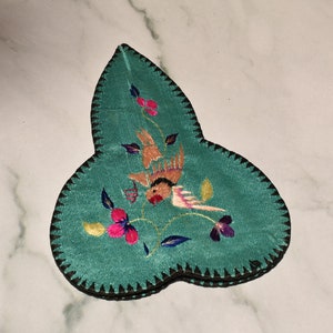 May include: A teal blue fabric leaf-shaped embroidered trinket box with a bird and floral design. The box has a black trim around the edges.