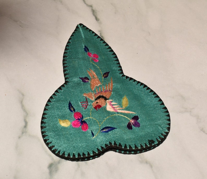 May include: A teal blue fabric leaf-shaped embroidered trinket box with a bird and floral design. The box has a black trim around the edges.