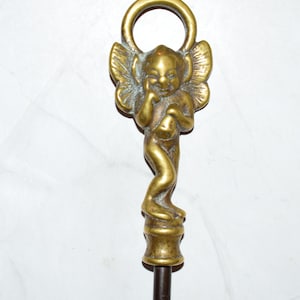 May include: A brass fireplace tool handle featuring a sculpted winged figure with a hand resting on its cheek. The figure stands atop a cylindrical base, and a circular ring is positioned directly above the head of the winged character.