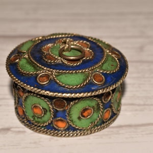 Op de afbeelding: Een kleine, ronde decoratieve doos met een scharnierend deksel. De doos heeft een blauw, groen en oranje emaille ontwerp met een zilverkleurige metalen rand. Het deksel heeft een kleine ringgreep. Voor het opbergen van kleine items.