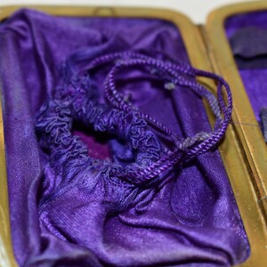 May include: An open antique case with a gold-coloured frame and a deep purple satin interior. A matching drawstring pouch sits inside, with a twisted cord. The case is open, revealing a small compartment.