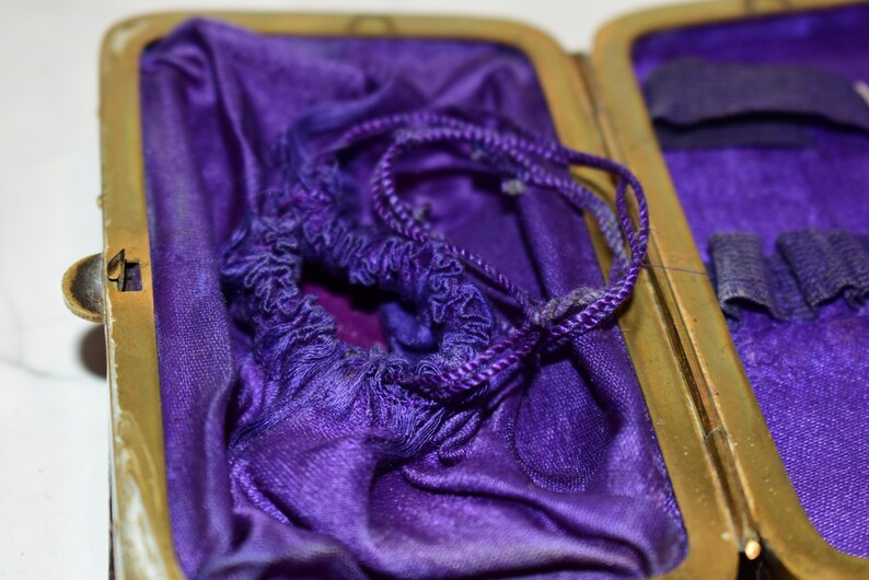 May include: An open antique case with a gold-coloured frame and a deep purple satin interior. A matching drawstring pouch sits inside, with a twisted cord. The case is open, revealing a small compartment.