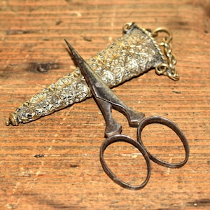 May include: Antique metal scissors with a decorative sheath. The scissors are dark gray with a pointed tip and rounded finger holes. The sheath is silver-colored with an ornate, embossed pattern and a small chain.
