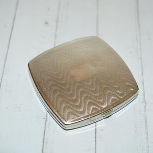 Op de afbeelding: Een zilverkleurige, vierkante compacte poederdoos met een gestructureerd, golvend patroon op het deksel. De compact heeft een rechthoekig accent in het midden en een dunne, zwarte rand.