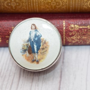 Op de afbeelding: Een ronde, zilverkleurige sieradendoos met een witte achtergrond en een kleurrijk beeld van een persoon in een blauwe jas en witte broek die in een veld staat.