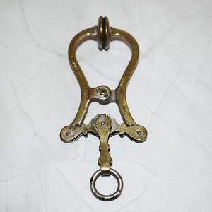 Op de afbeelding: Antieke messingkleurige metalen tool. Het heeft een hartvormige bovenkant, een scharnierend middengedeelte en een kleine ring aan de onderkant. De tool heeft decoratieve details en vertoont tekenen van ouderdom, met een aangetaste uitstraling van het metaal.