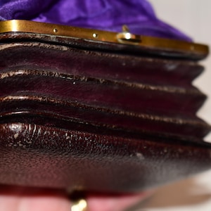 May include: Close-up of a vintage, dark brown leather purse with a gold-coloured clasp. The purse has a pleated design and a purple satin lining. The leather shows signs of wear, adding to its antique charm.