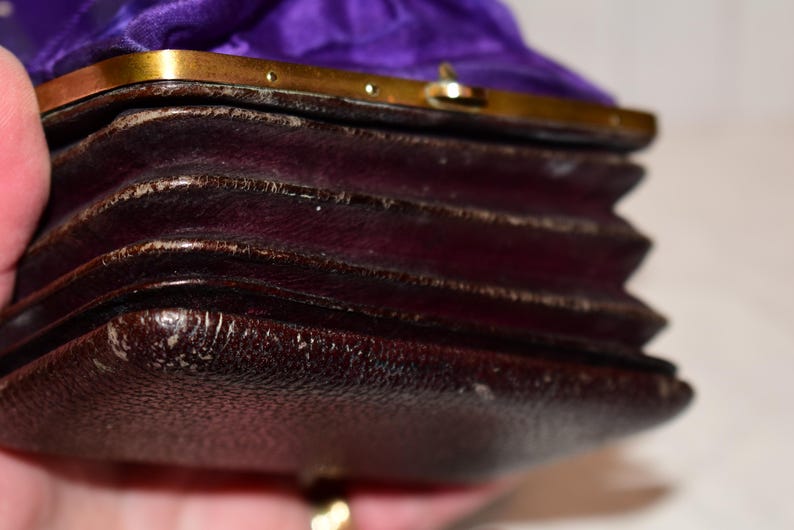 May include: Close-up of a vintage, dark brown leather purse with a gold-coloured clasp. The purse has a pleated design and a purple satin lining. The leather shows signs of wear, adding to its antique charm.