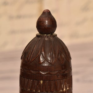 May include: A dark brown wooden finial with intricate carvings. The top of the finial is shaped like a teardrop and the bottom has a series of bands.
